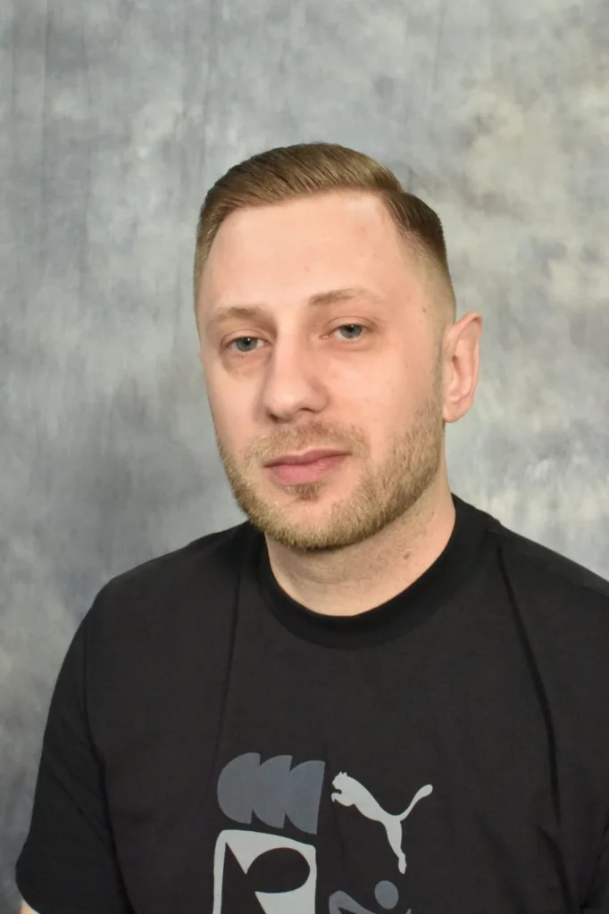 Amel Melkic vor neutralem Studiohintergrund, trägt ein schwarzes T-Shirt mit dezentem Motiv und blickt ruhig in die Kamera.