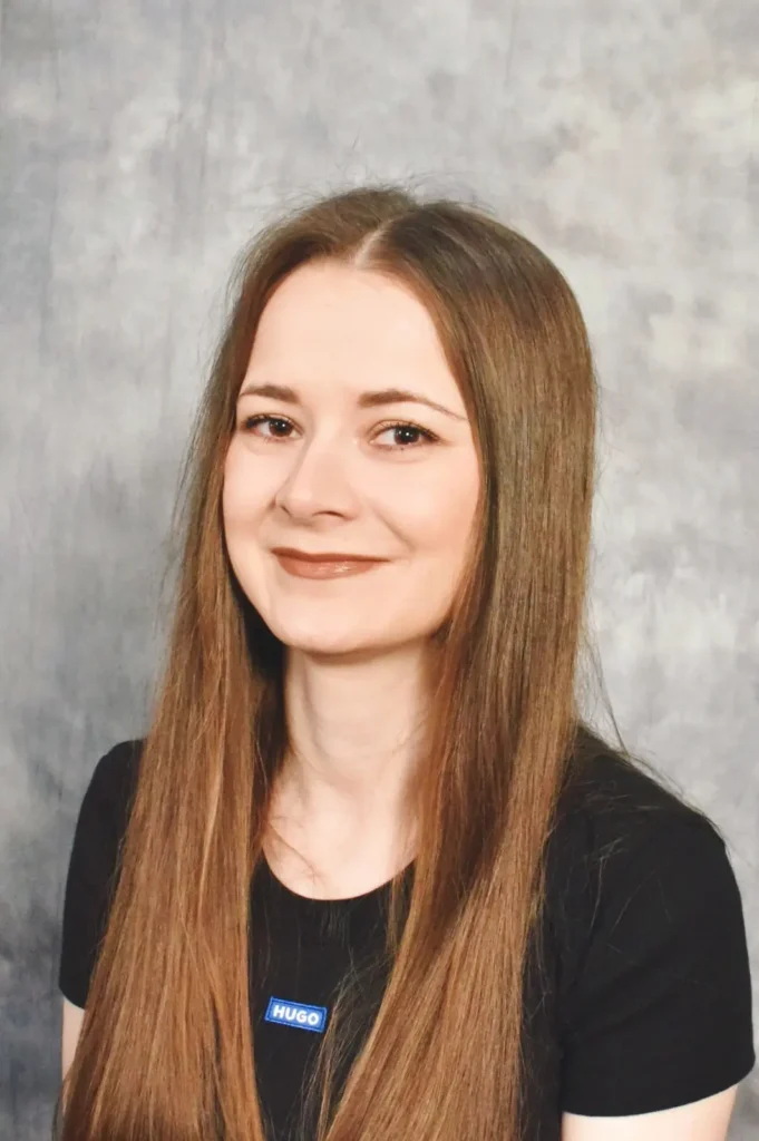 Anela lächelt in die Kamera, mit langen braunen Haaren vor neutralem Studiohintergrund.